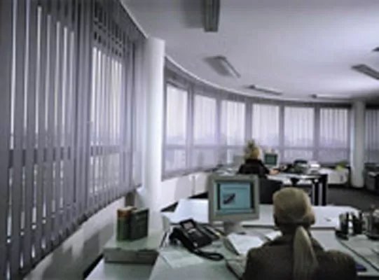 Graue Vertikaljalousien in einem Büroraum vor einer langen gebogenen Fensterfront.