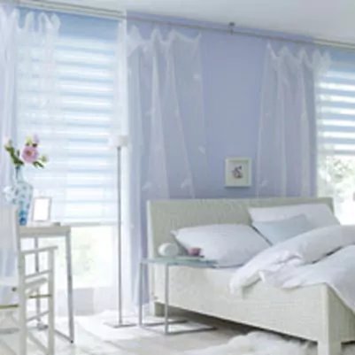 Hellblaue Doppelrollos an den beiden großen Fenstern eines Schlafzimmers.